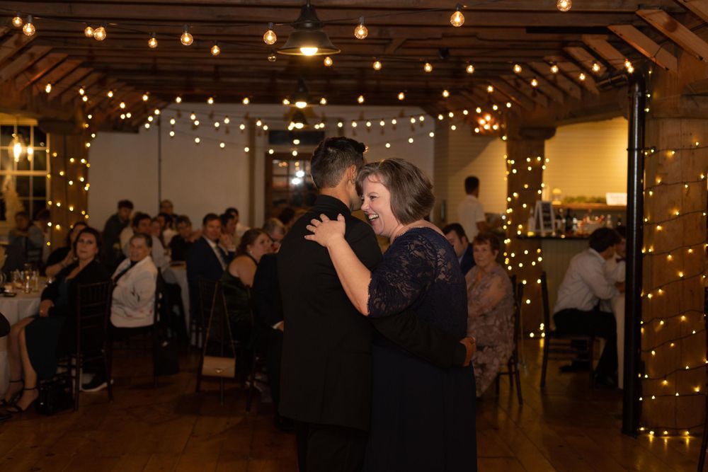Couples slow dancing at a wedding reception under warm string lights in a rustic venue.