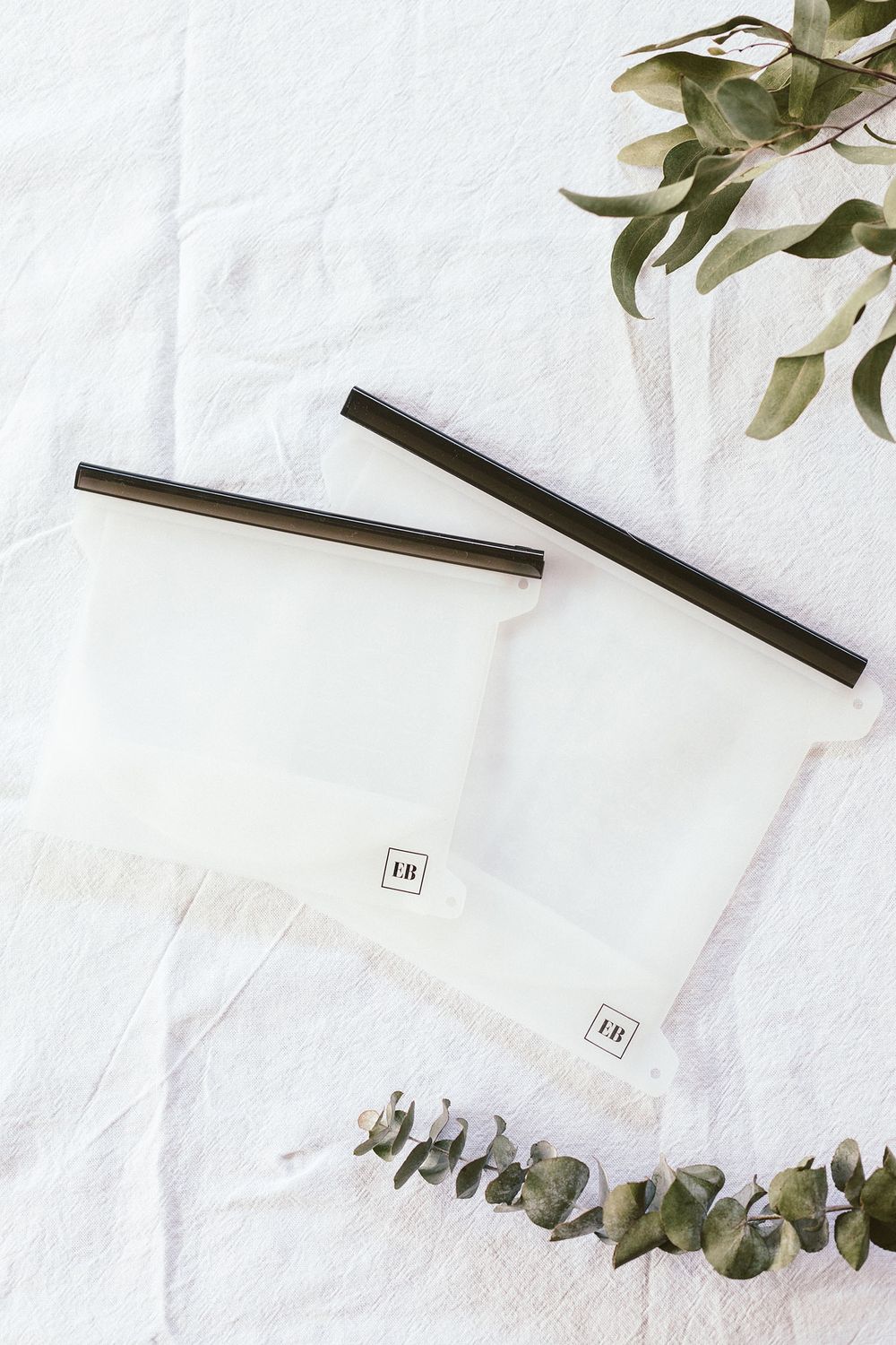 Reusable silicone storage bags with black sealing strips displayed with eucalyptus branches on white surface.