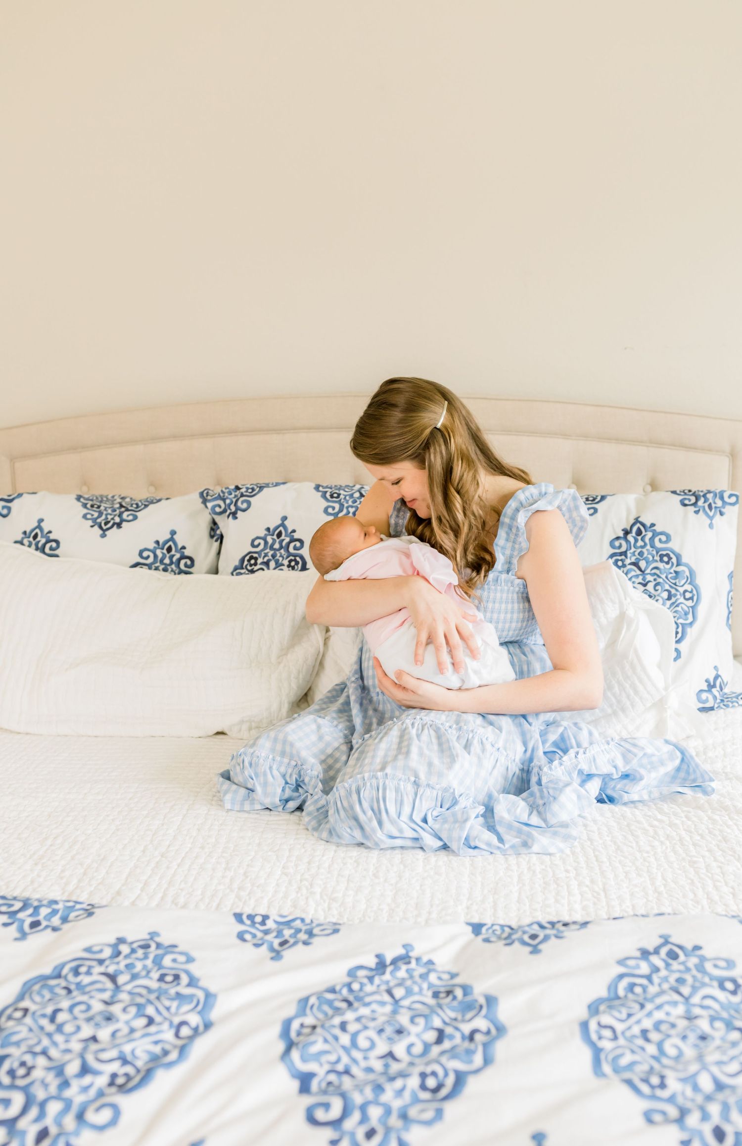 Mother in light blue dress cradles newborn baby on bed with decorative bedding.