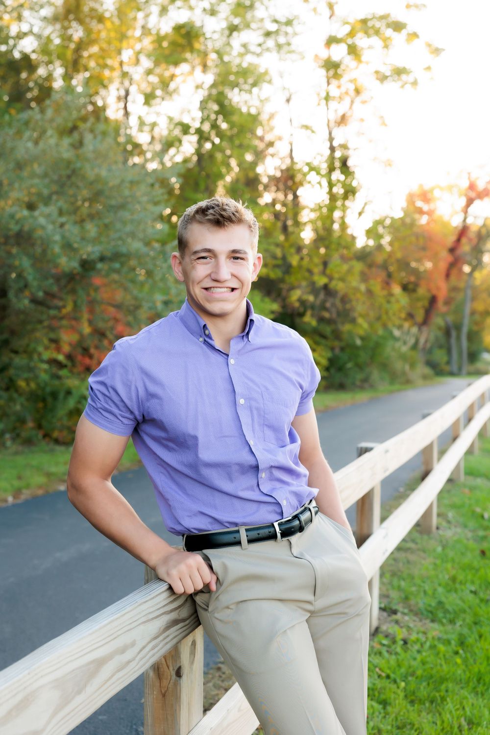 Person in purple button-down shirt and khakis poses casually by a wooden railing outdoors.