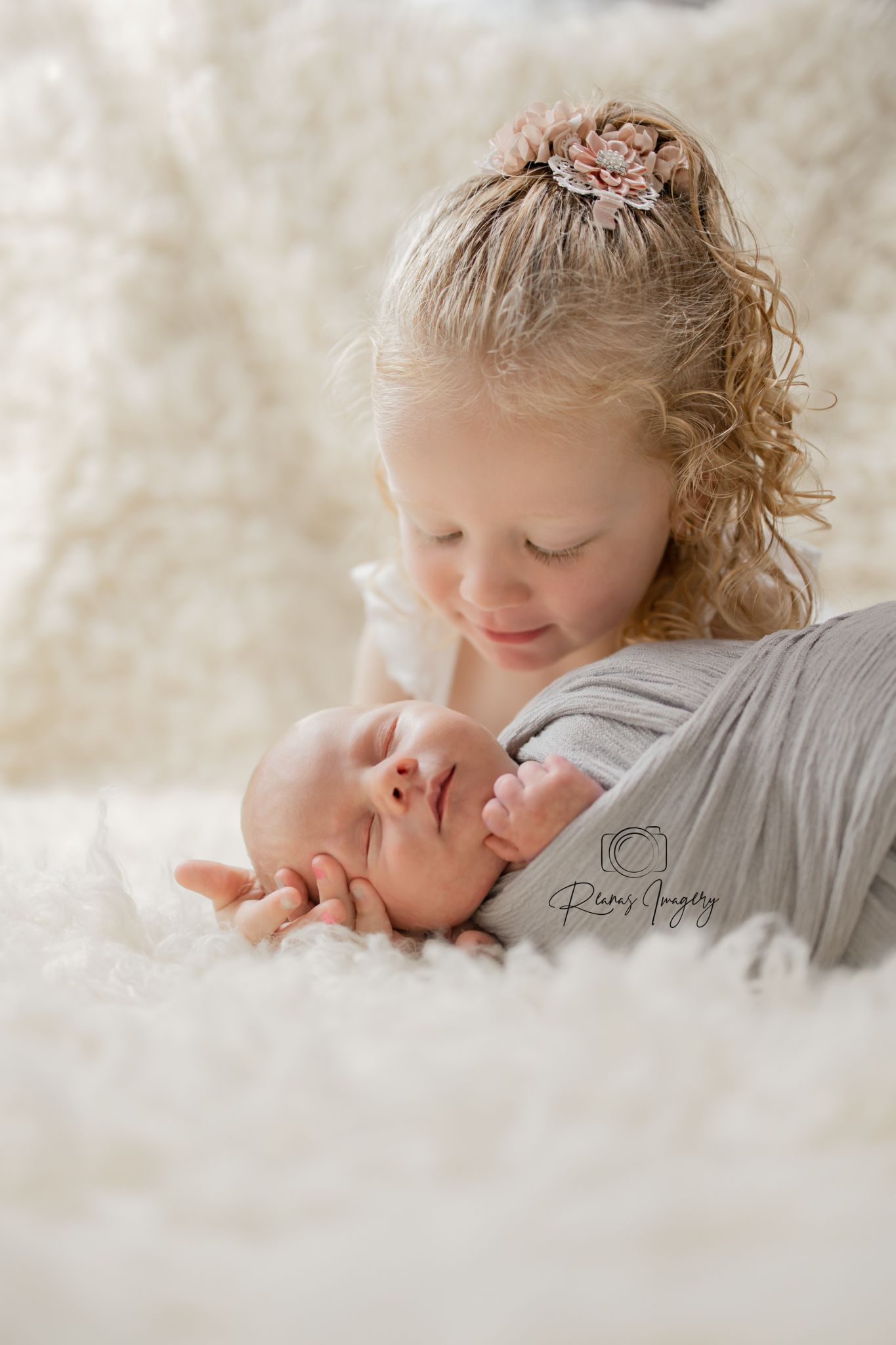 Curly-haired big sister gently cradles her smiling newborn sibling wrapped in a gray swaddle.