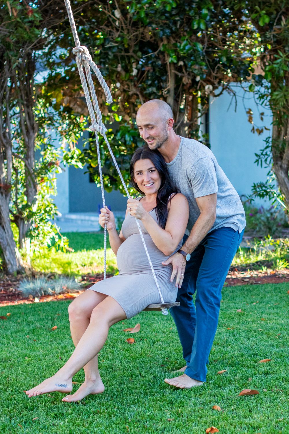 Smiling couple posing outdoors with pregnant woman sitting on rope swing in grey maternity dress.