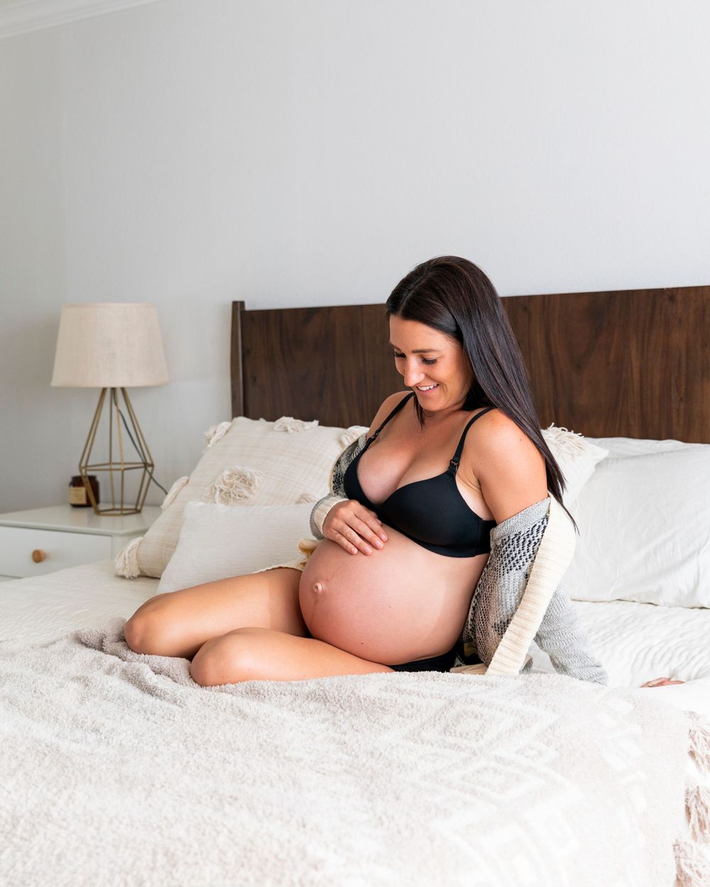 In-home maternity photoshoot with Pregnant brunette woman sitting on bed in black bra cradling bare baby bump during maternity shoot.