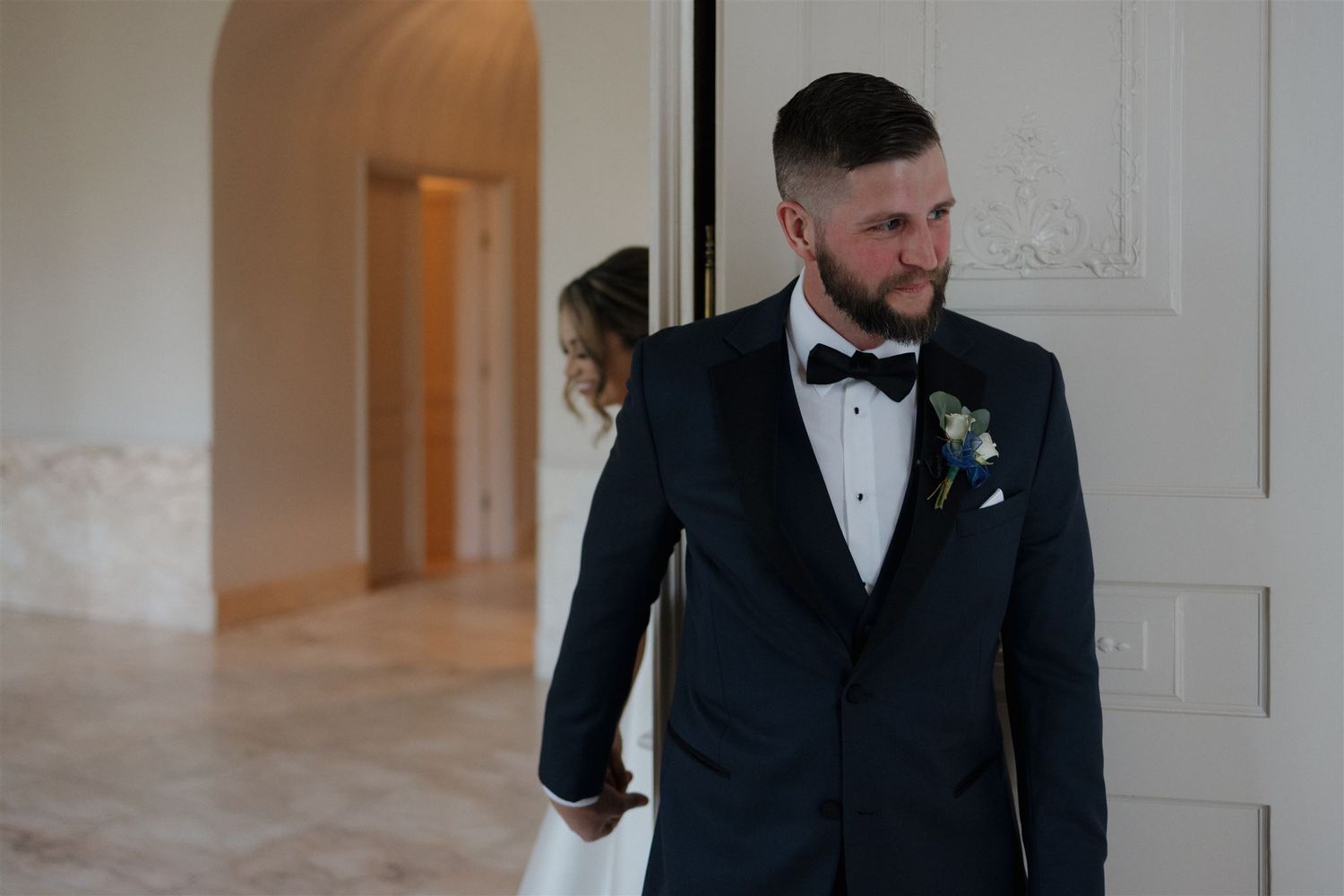 A groom in a classic black tuxedo and bowtie stands by a white doorway at a formal wedding venue.