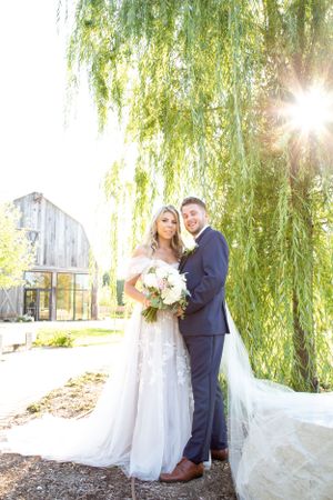 Sunlit portrait of newlyweds under a weeping willow tree with bride holding white bouquet.