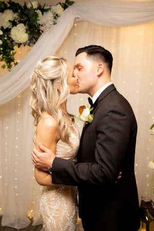 Romantic kiss between newlyweds against a backdrop of white draping and twinkling lights.