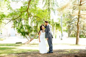 Couple in wedding attire embracing under a weeping willow tree in a sunlit park.