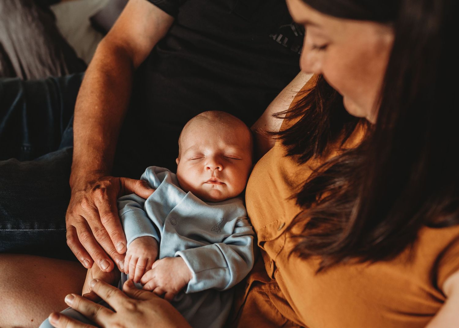 A sleeping infant in light blue clothing rests peacefully on a couch with parents.