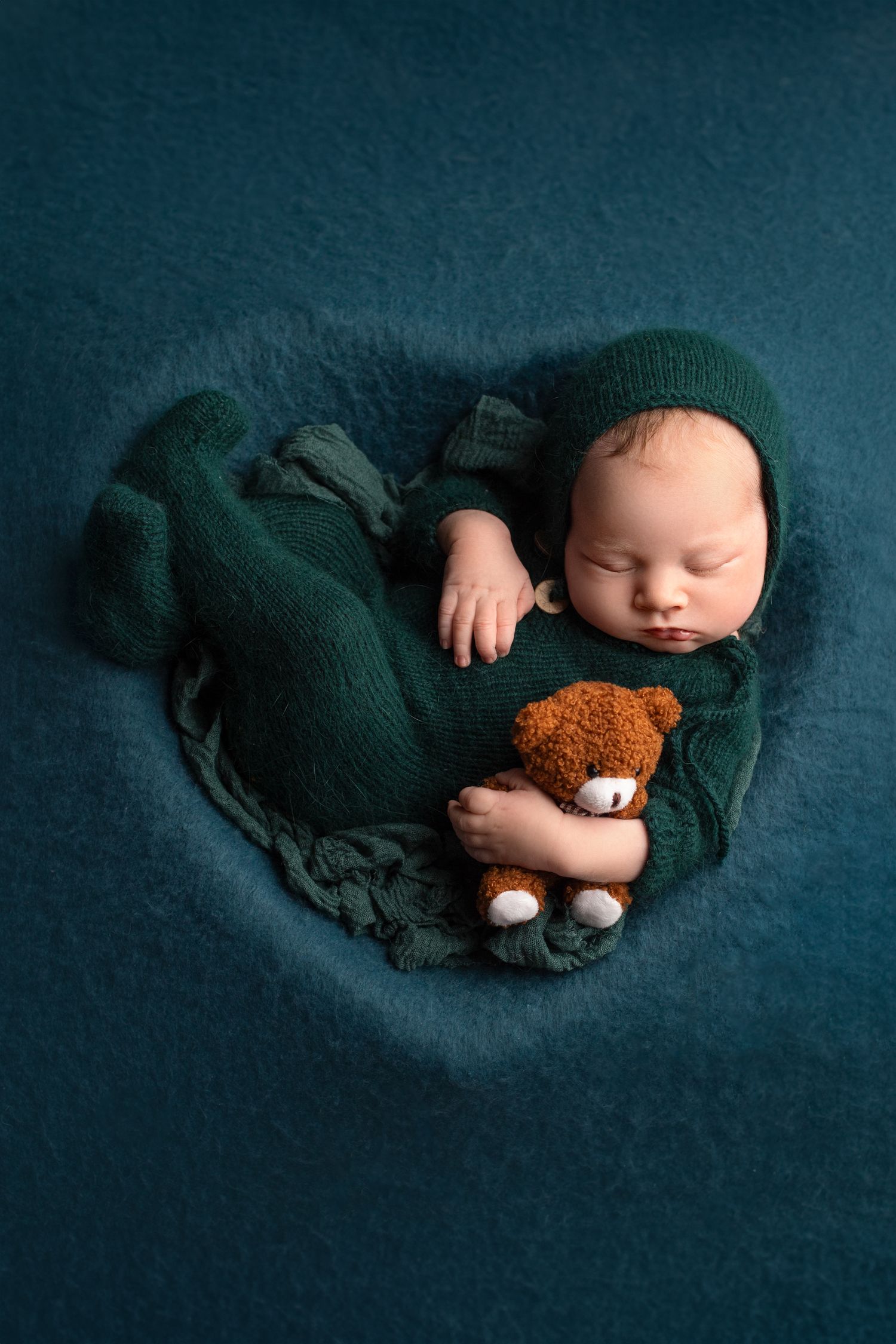 Sleeping infant wrapped in green blanket holding small teddy bear on teal background. springdield lakes studio fine art