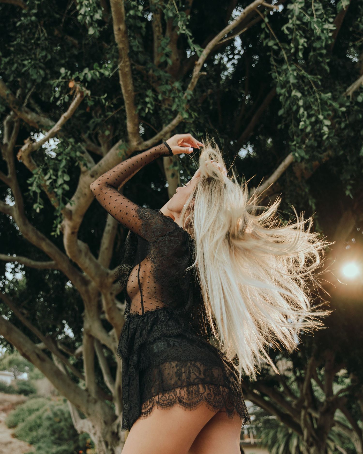 Woman in black lace dress dancing with flowing blonde hair against sunlit trees.