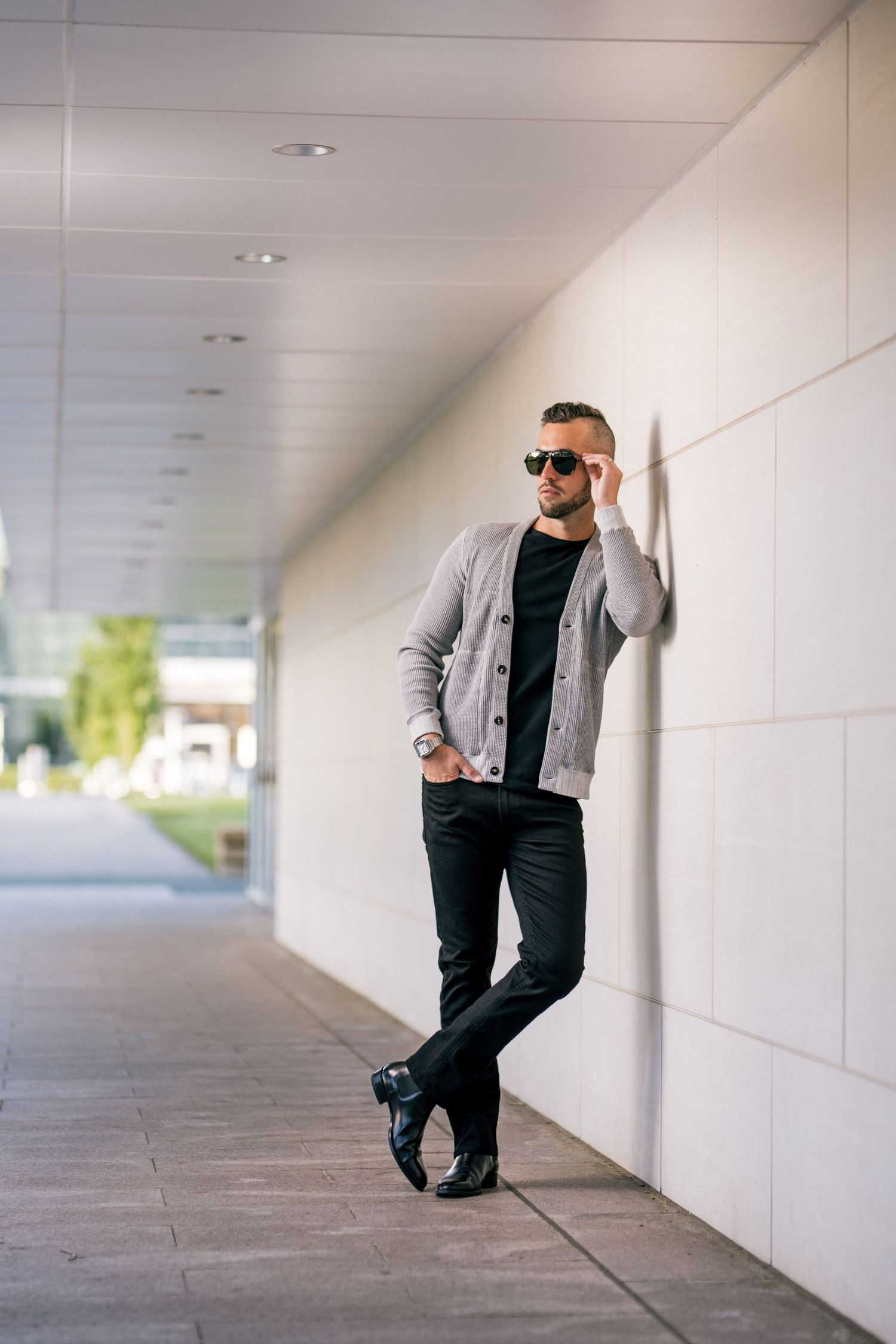 Casual portrait of person in grey jacket and sunglasses leaning against white wall.