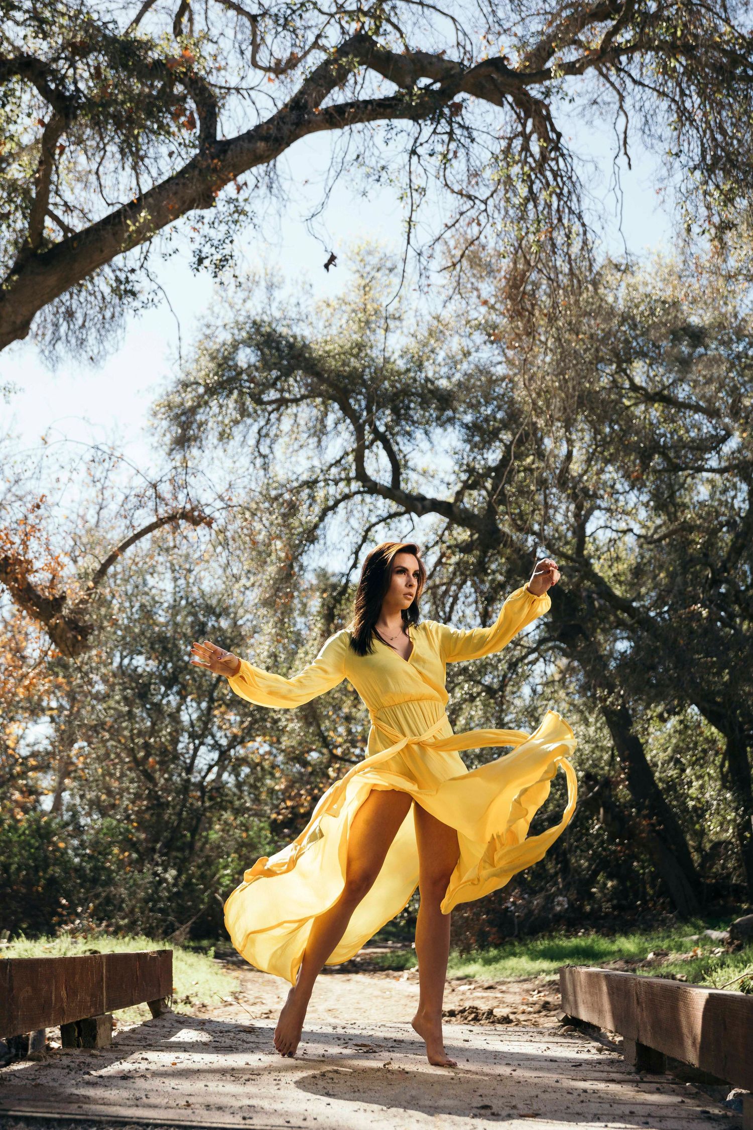 Woman in flowing yellow dress twirling outdoors under tree branches.