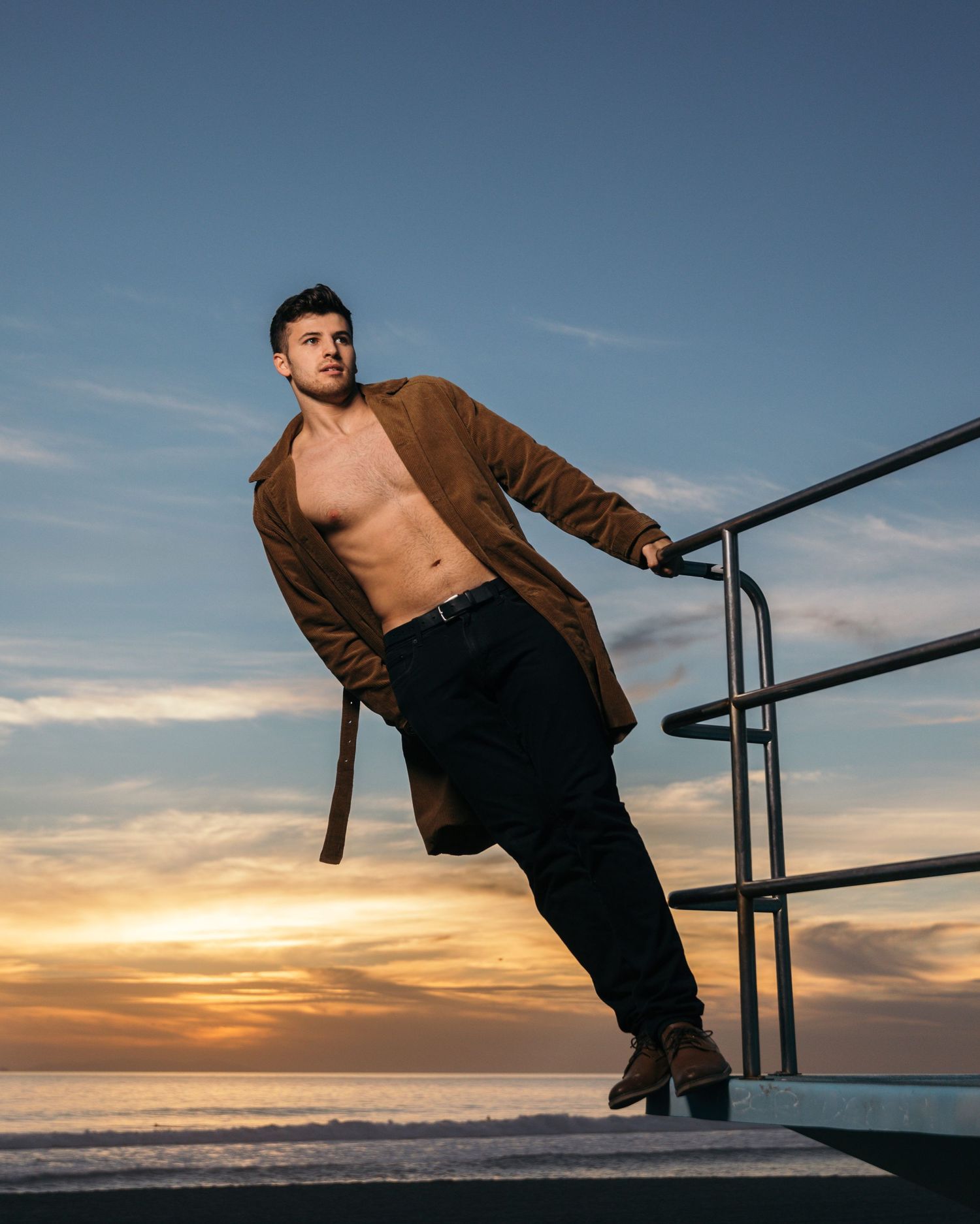 Athletic man in brown jacket leaning on railing during sunset at beach.