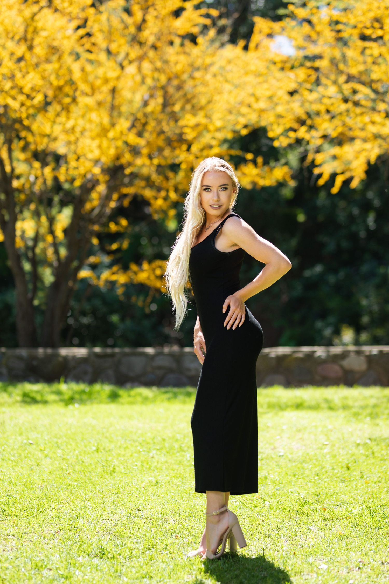 Woman in black dress poses on lawn with yellow autumn trees in background.