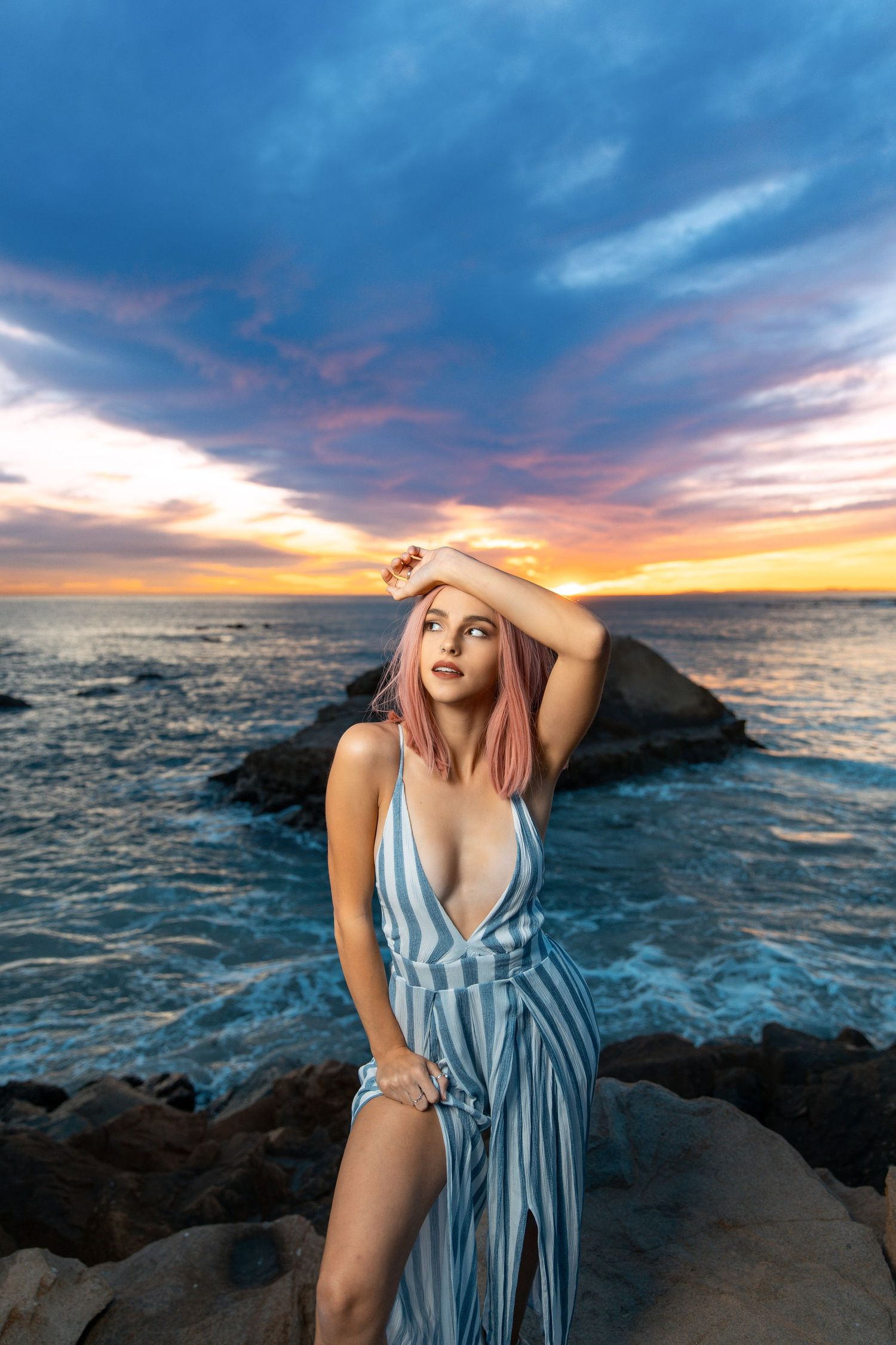 Woman in striped blue dress poses against stunning ocean sunset.