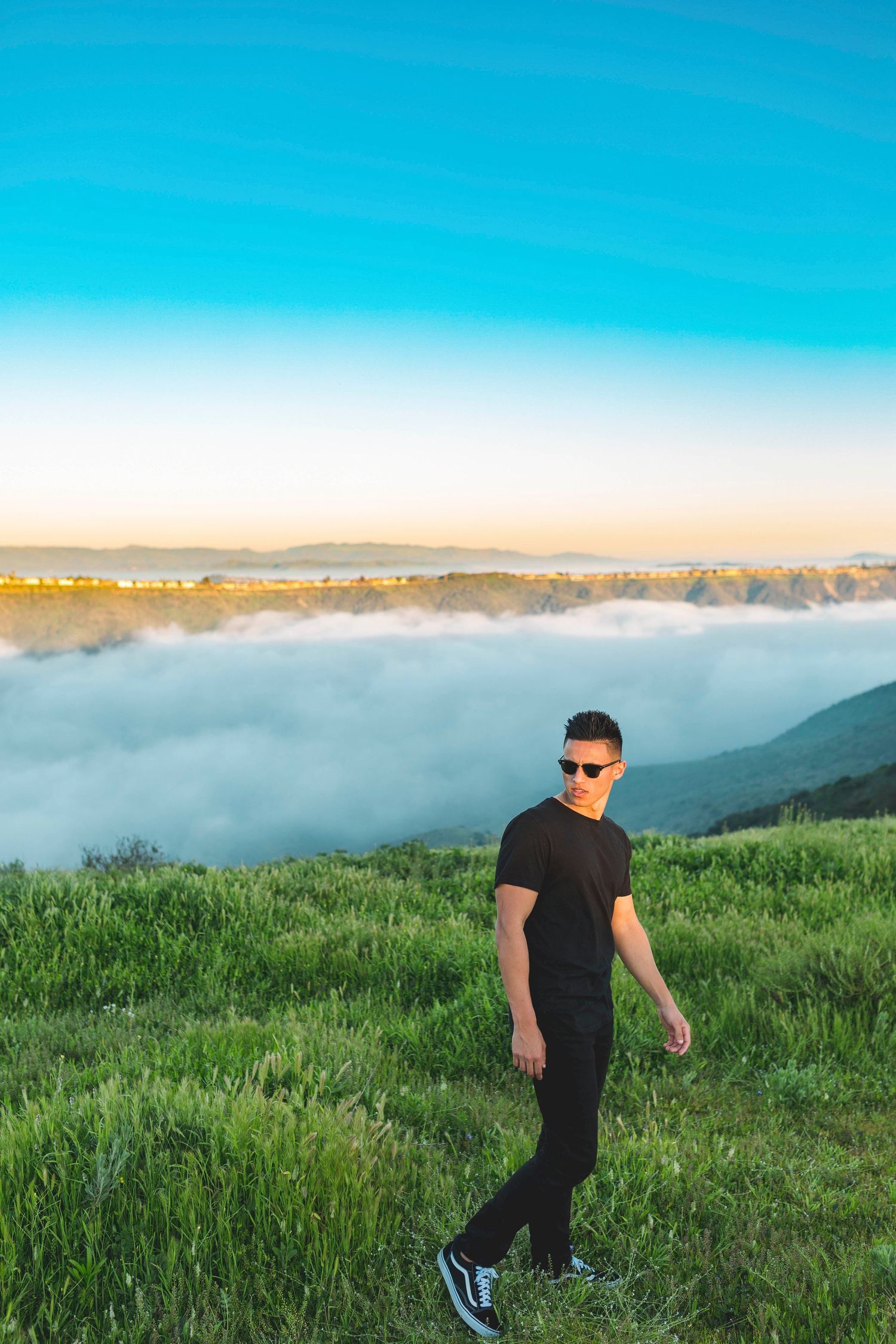 Person in black outfit walking on grassy hillside above fog layer at sunset.