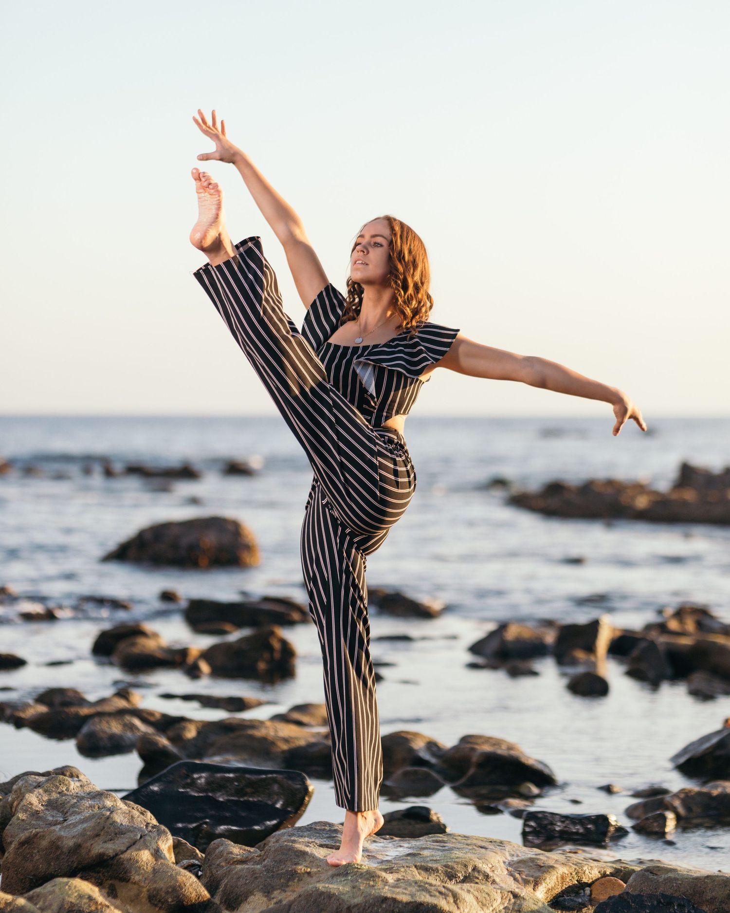 Dancer in striped jumpsuit performs graceful pose on rocky beach at sunset.