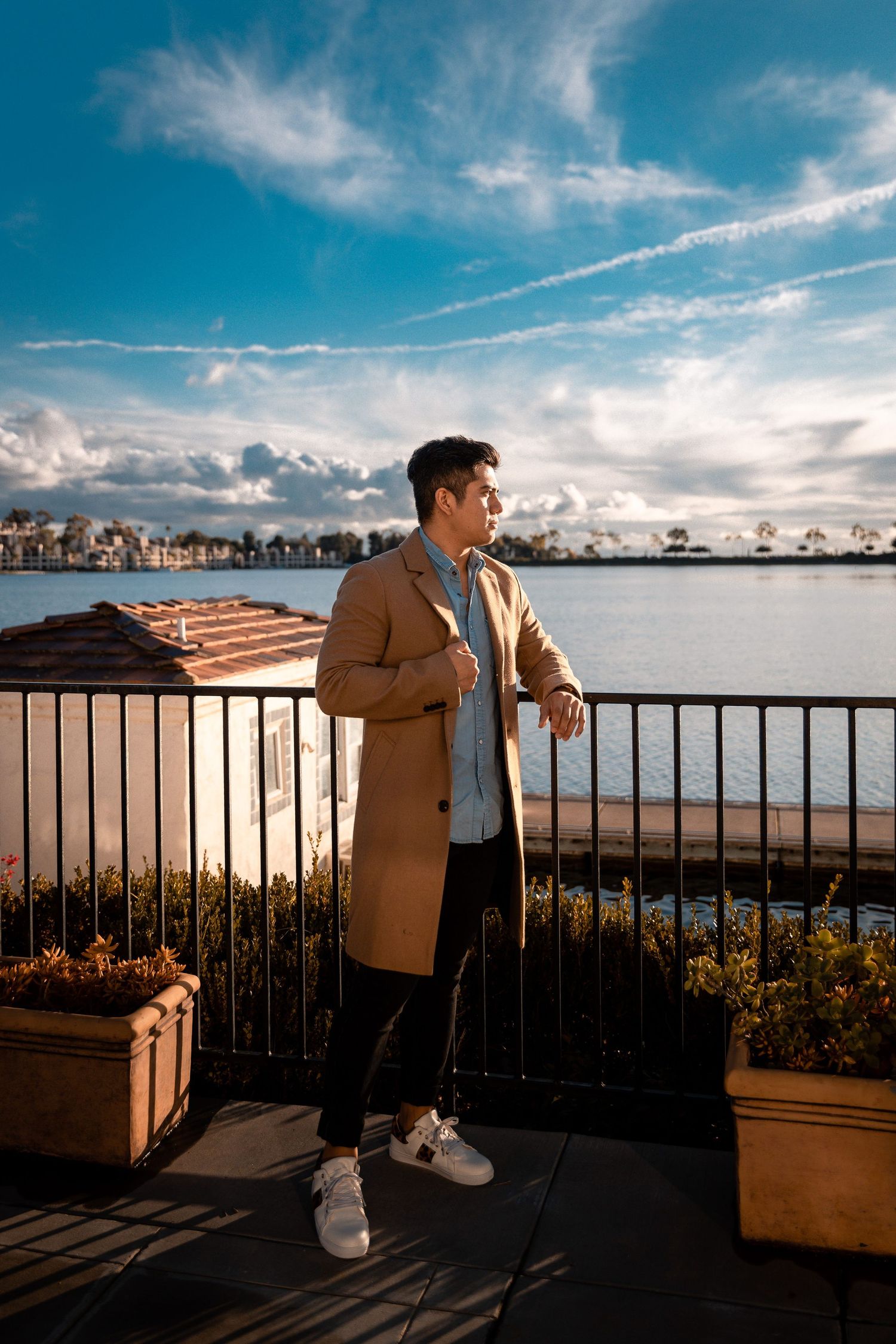 Man in tan coat poses on waterfront balcony against harbor backdrop.