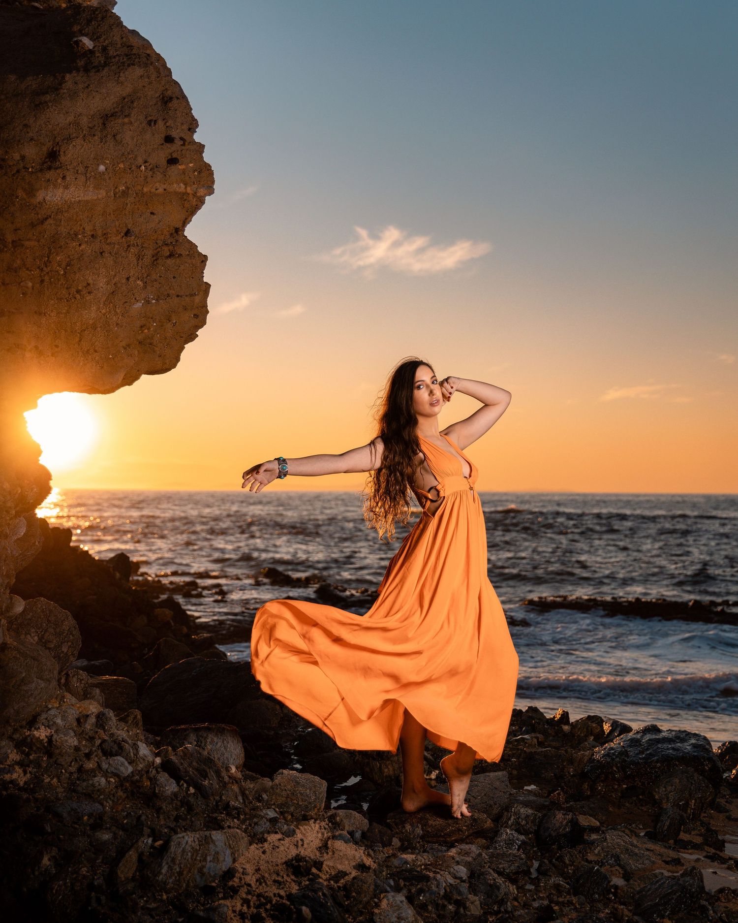 Orange flowing dress catches wind as figure poses dramatically on rocky beach during sunset.