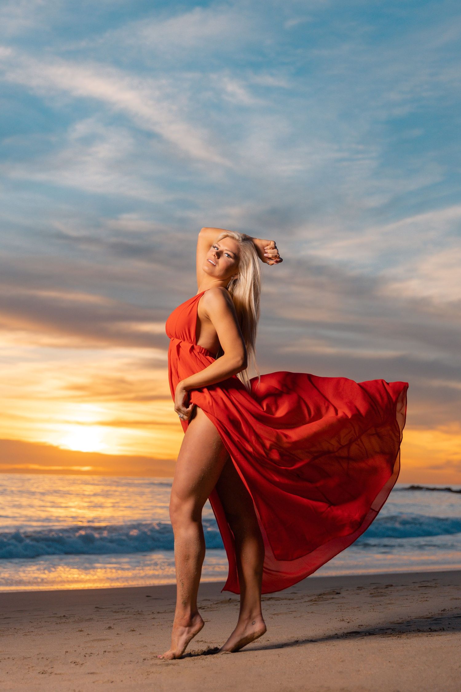 Flowing red dress catches wind during beach sunset photoshoot.