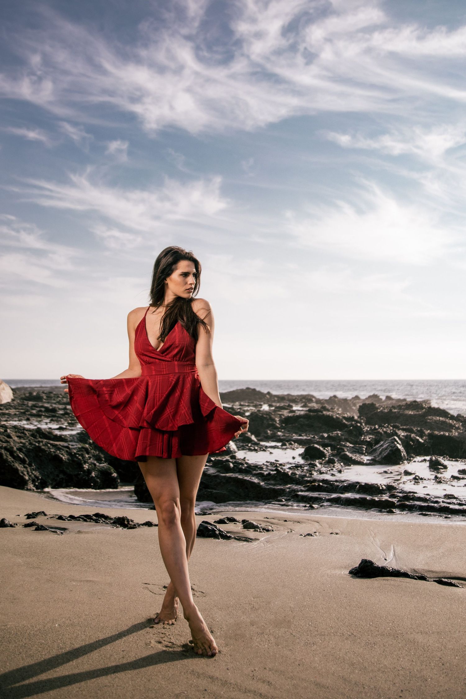 Woman in flowing red dress poses dramatically on a beach with rocky coastline and cloudy sky.