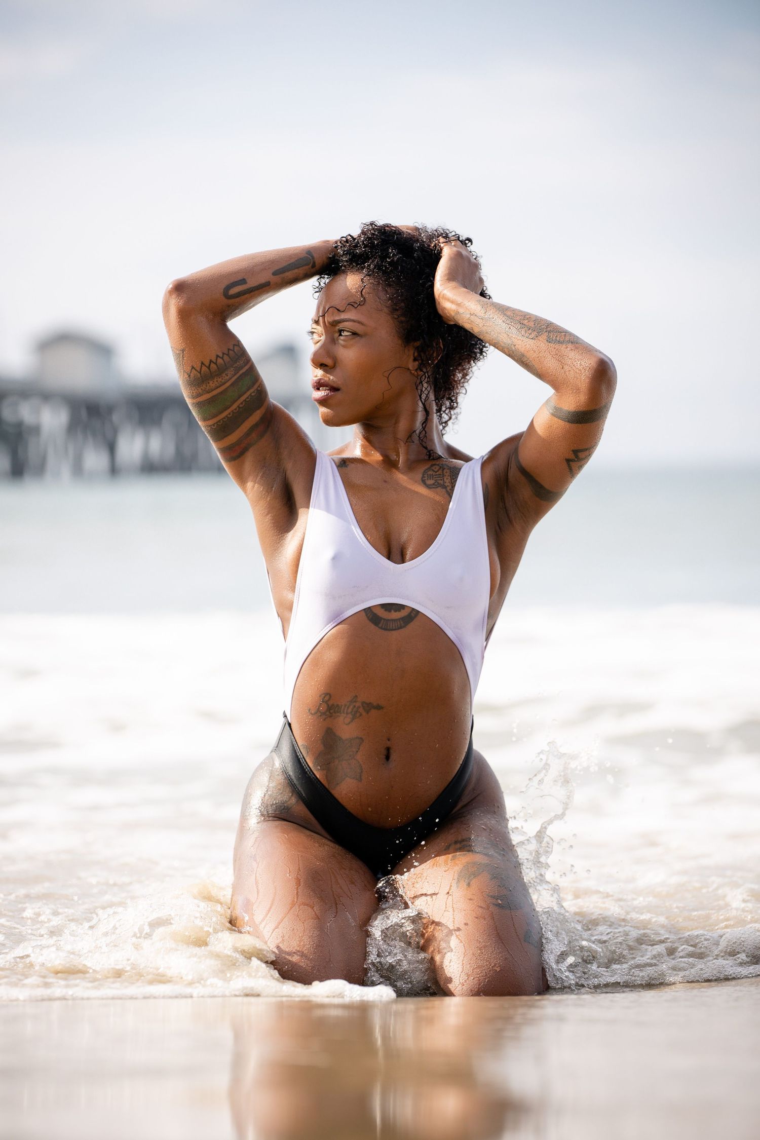 Athletic figure in white swimsuit kneeling in ocean waves at beach.