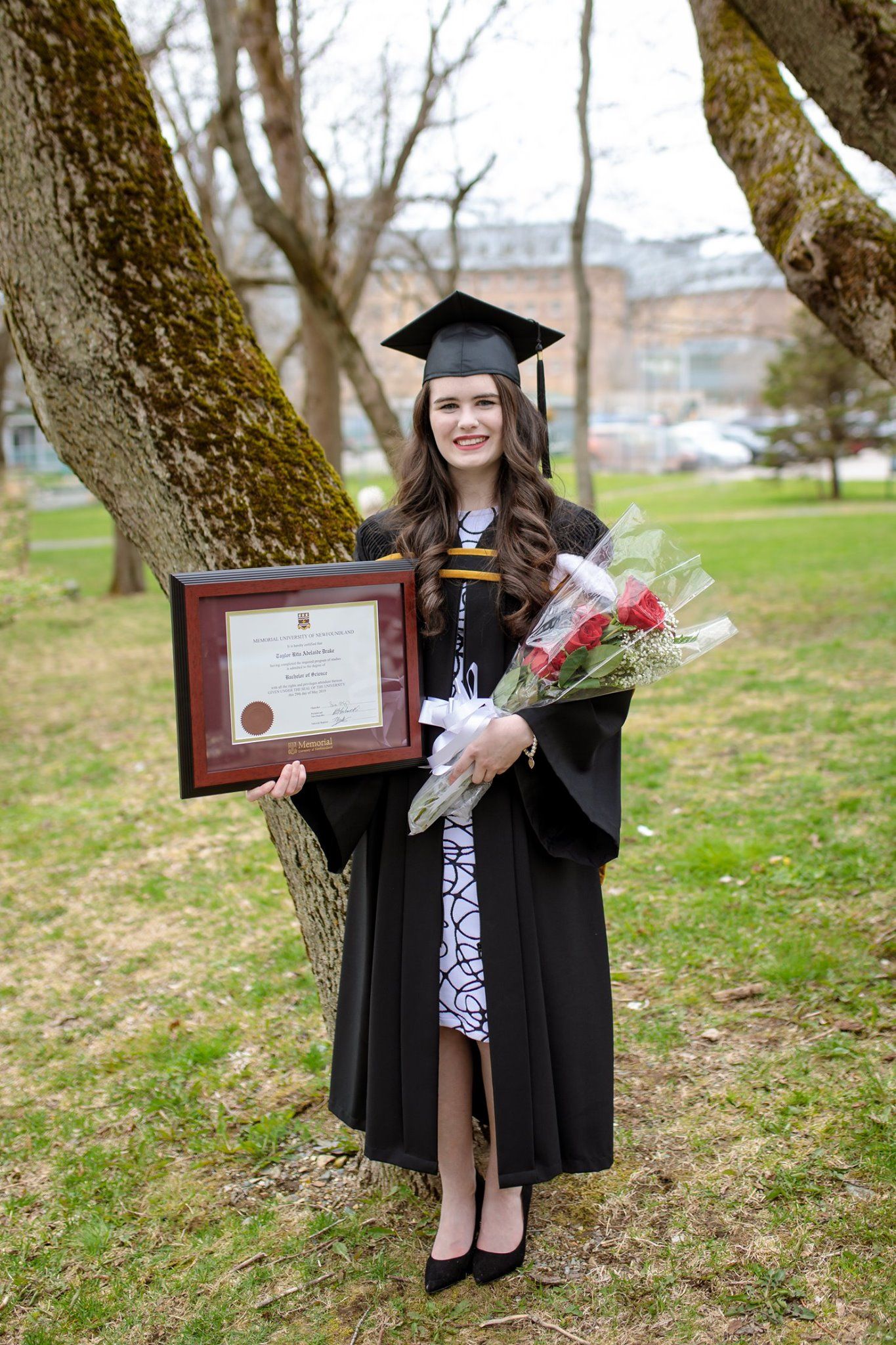 A graduate in cap and gown holds a diploma and flowers while posing by a tree.