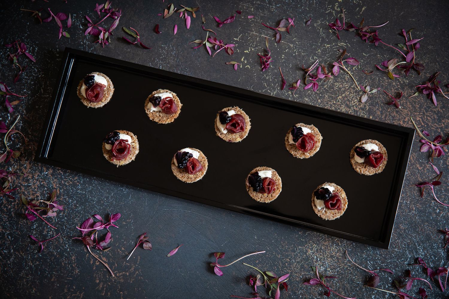 Black plate displaying elegant mini desserts garnished with purple flower petals on dark background.