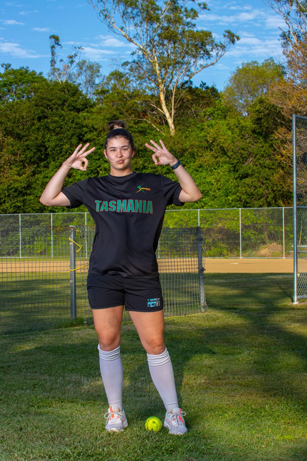 Athlete in Tasmania uniform makes peace signs while posing with tennis ball on sports court.