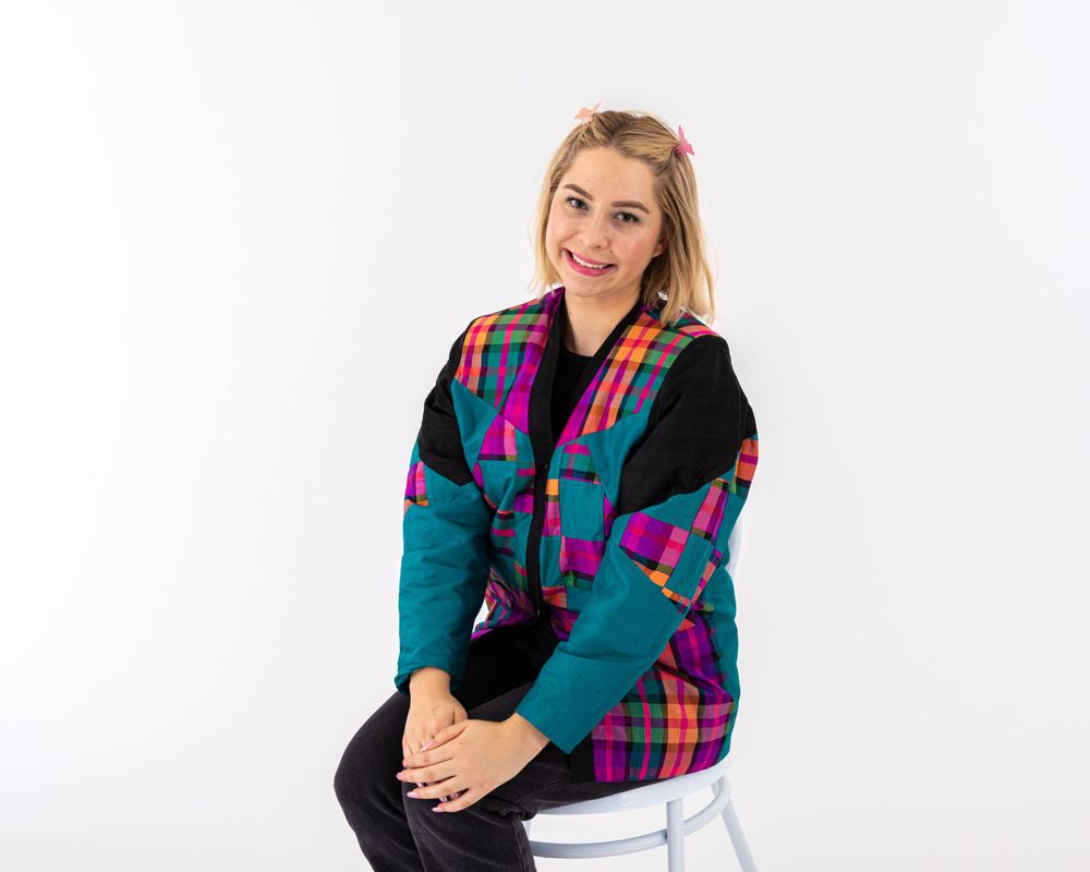 A person wearing a colorful plaid and teal jacket poses on a white stool.