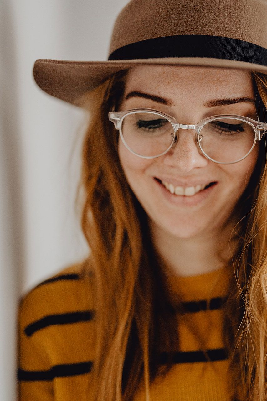 A close-up portrait with a brown fedora hat, clear glasses, and striped mustard yellow sweater.