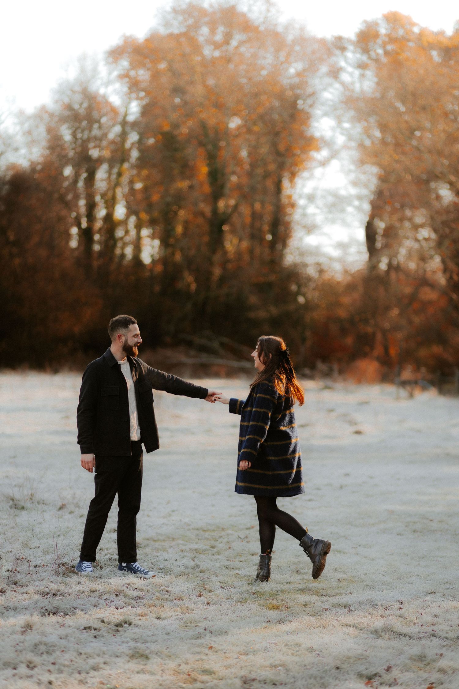 A couple dances playfully in a frosty field surrounded by autumn trees.