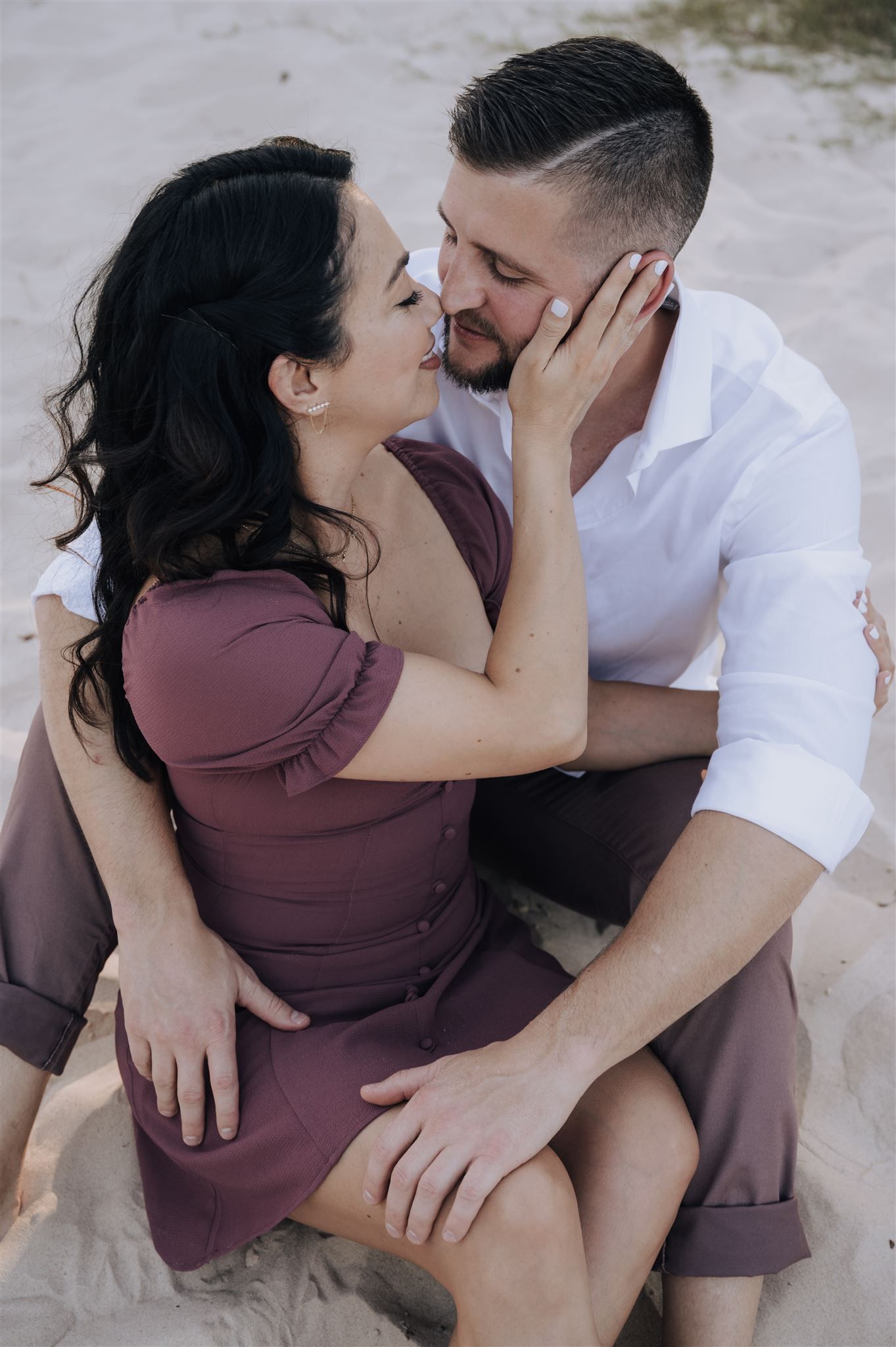 Romantic couple in burgundy and white outfits sharing tender moment while sitting on sandy beach.