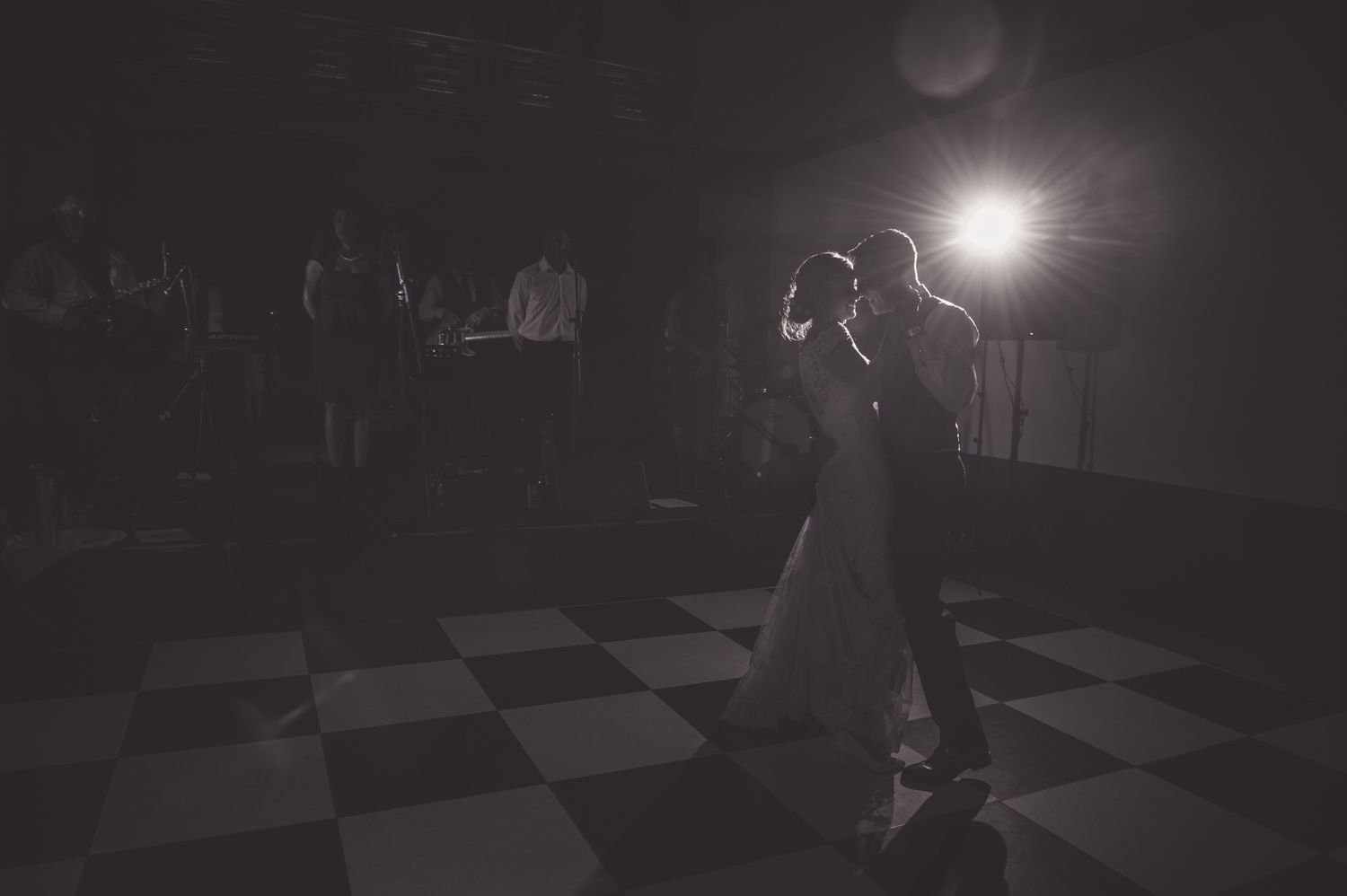 Couple shares first dance in dramatic spotlight on checkered dance floor.