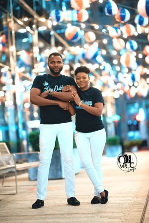 Coordinated couple in matching black shirts and white pants pose against illuminated backdrop.