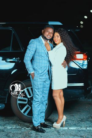 Stylish couple in light blue suit and white dress pose by luxury SUV.