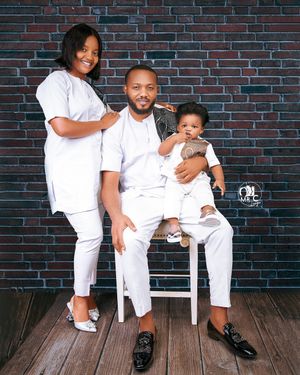 Family of three dressed in all white outfits poses together against a brick wall background.