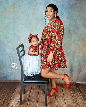 Mother and daughter wearing matching African print dresses pose on wooden chair against blue backdrop.