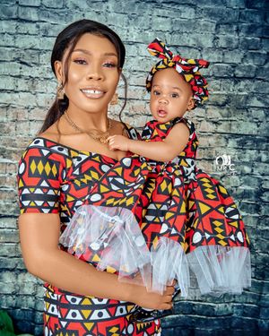 Mother and baby in matching colorful African print outfits with white tulle details pose against brick wall.