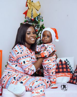 Mother and baby in matching holiday pajamas pose by Christmas tree with festive decorations.