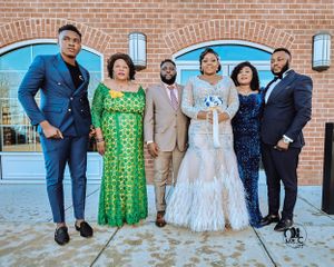 Formal group photo of wedding party in elegant attire standing outside brick building.