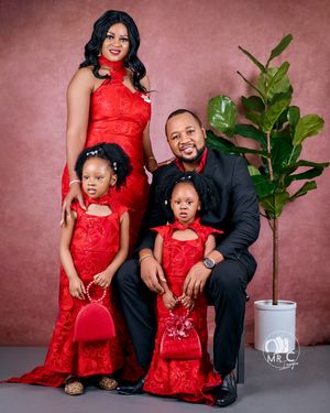 Family portrait in coordinated red formal wear with black suit against pink backdrop.