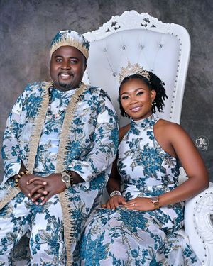 A couple in matching floral outfits and crowns pose on an ornate white throne chair.