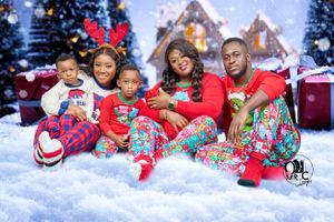 Family wearing matching festive Christmas pajamas poses together on snowy backdrop with gingerbread house decorations.