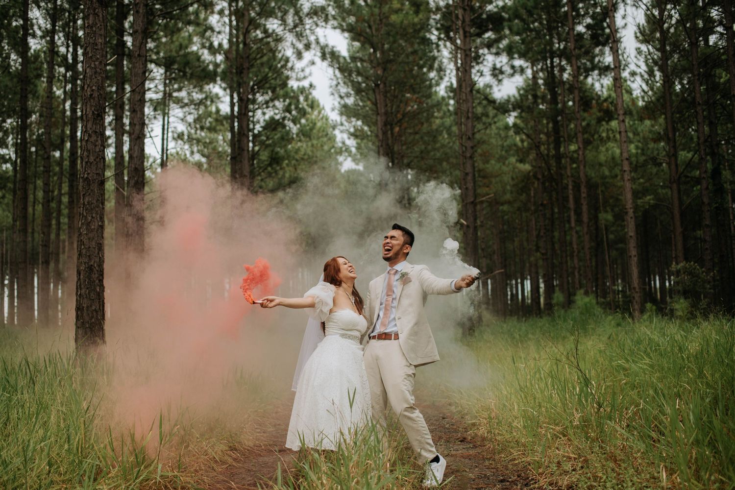 A couple celebrates with colorful smoke bombs in a pine forest setting during their wedding photoshoot.