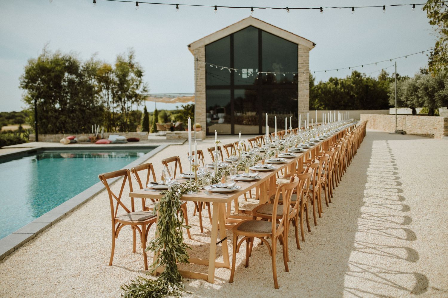 Rustic wooden chairs line a long dining table beside a modern pool with glass-walled building in the background.