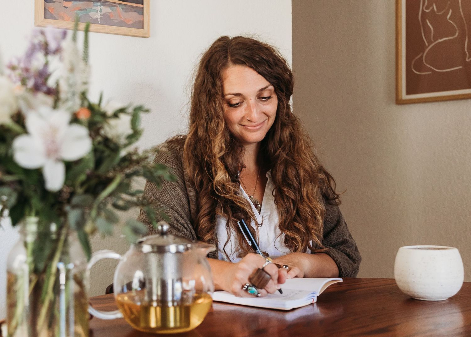 Someone journaling at a wooden table with flowers and tea setup in warm interior lighting.