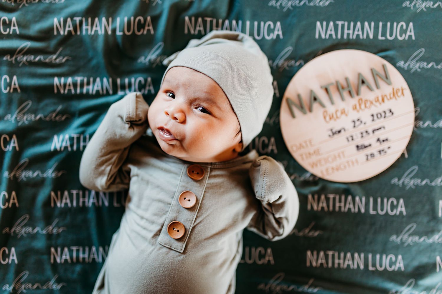 Newborn baby in gray outfit lies on custom name-patterned background.