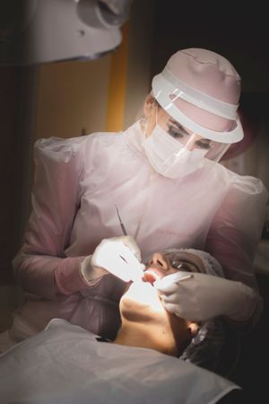 Healthcare provider in pink uniform conducting dental work under focused lighting.