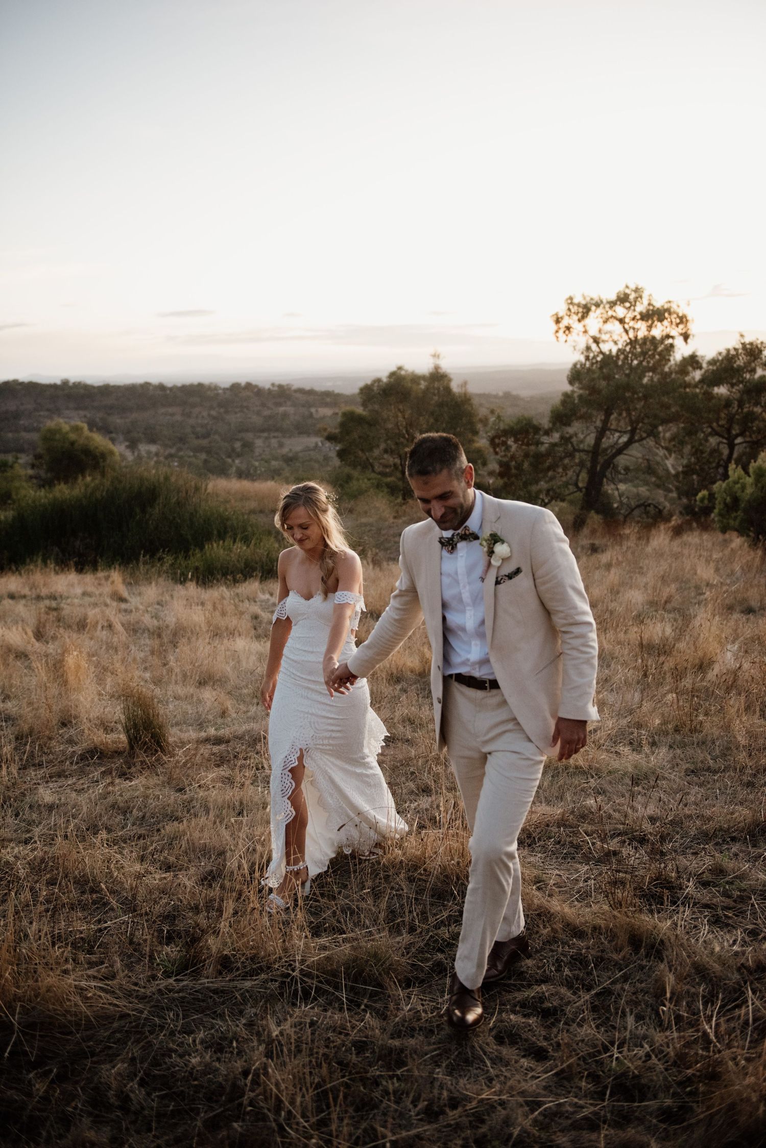 A stylish couple in white attire walking through golden grass at sunset.